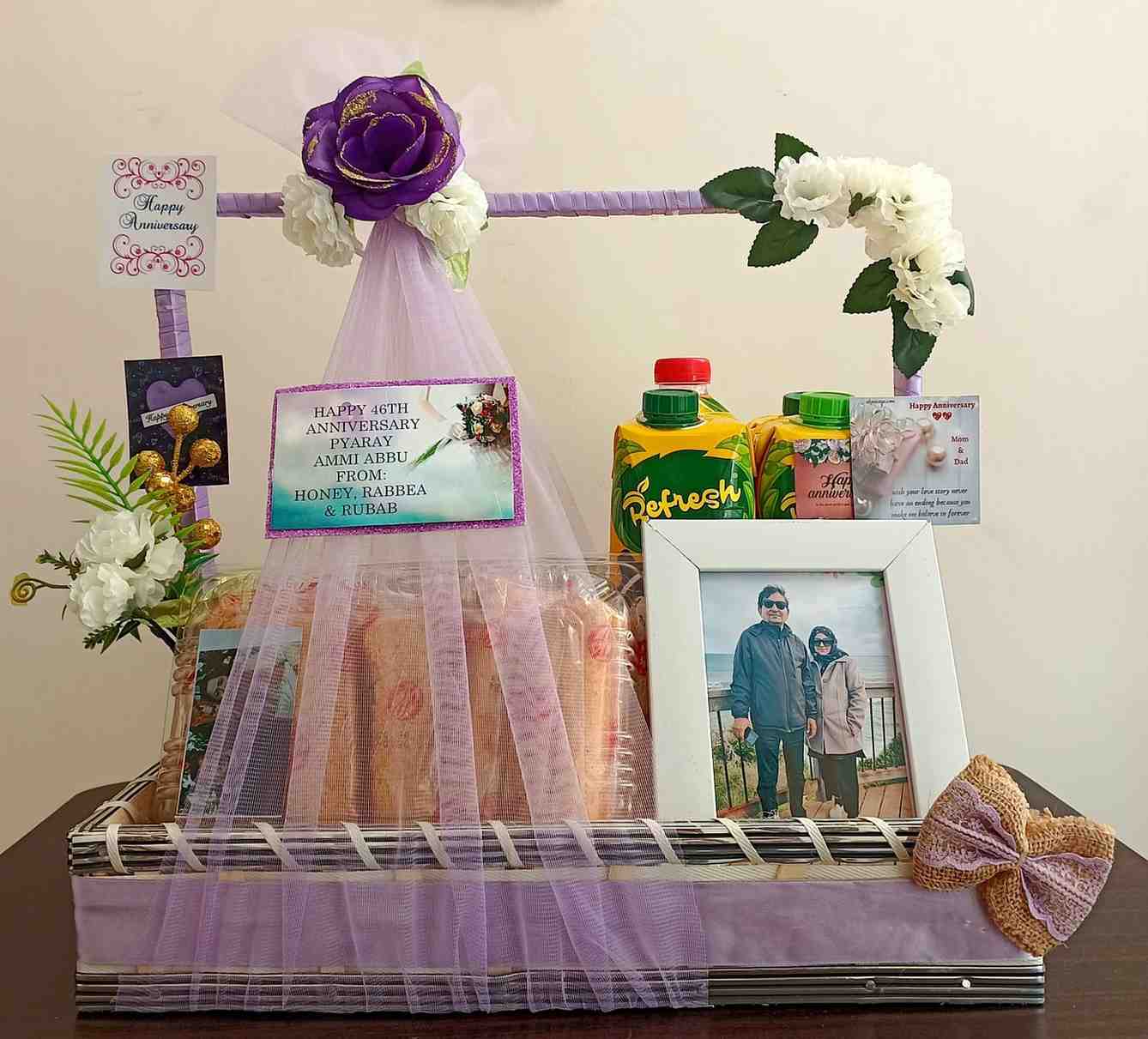 Purple-themed anniversary gift basket with personalised messages, a framed photo, juice bottles, and elegant floral decorations, perfect for a special celebration.