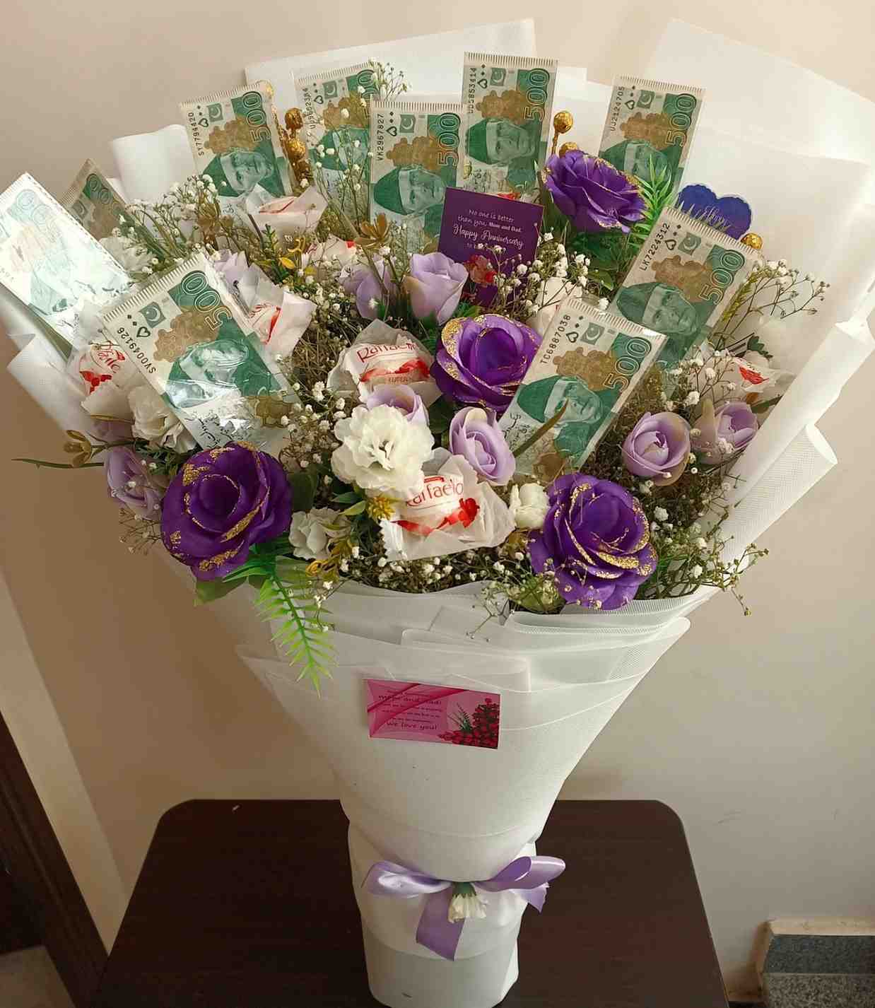 Luxurious bouquet with purple and white flowers, Pakistani currency notes, Raffaello chocolates, and elegant white wrapping tied with a purple ribbon.